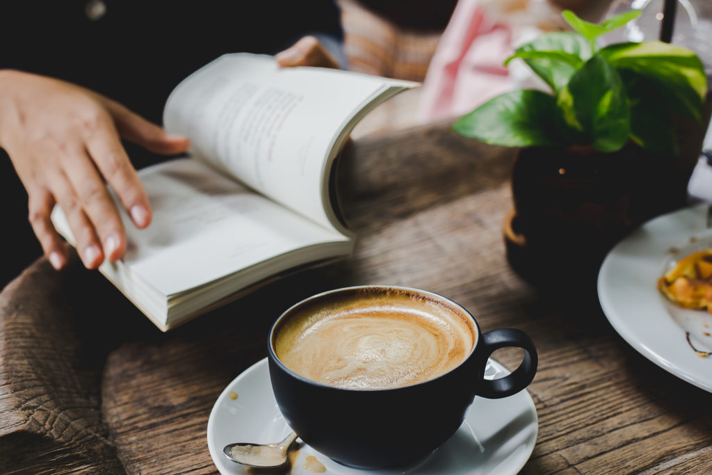 Stirred coffee cup on a saucer with a used spoon in front of a person reading a book at a wooden cafe table